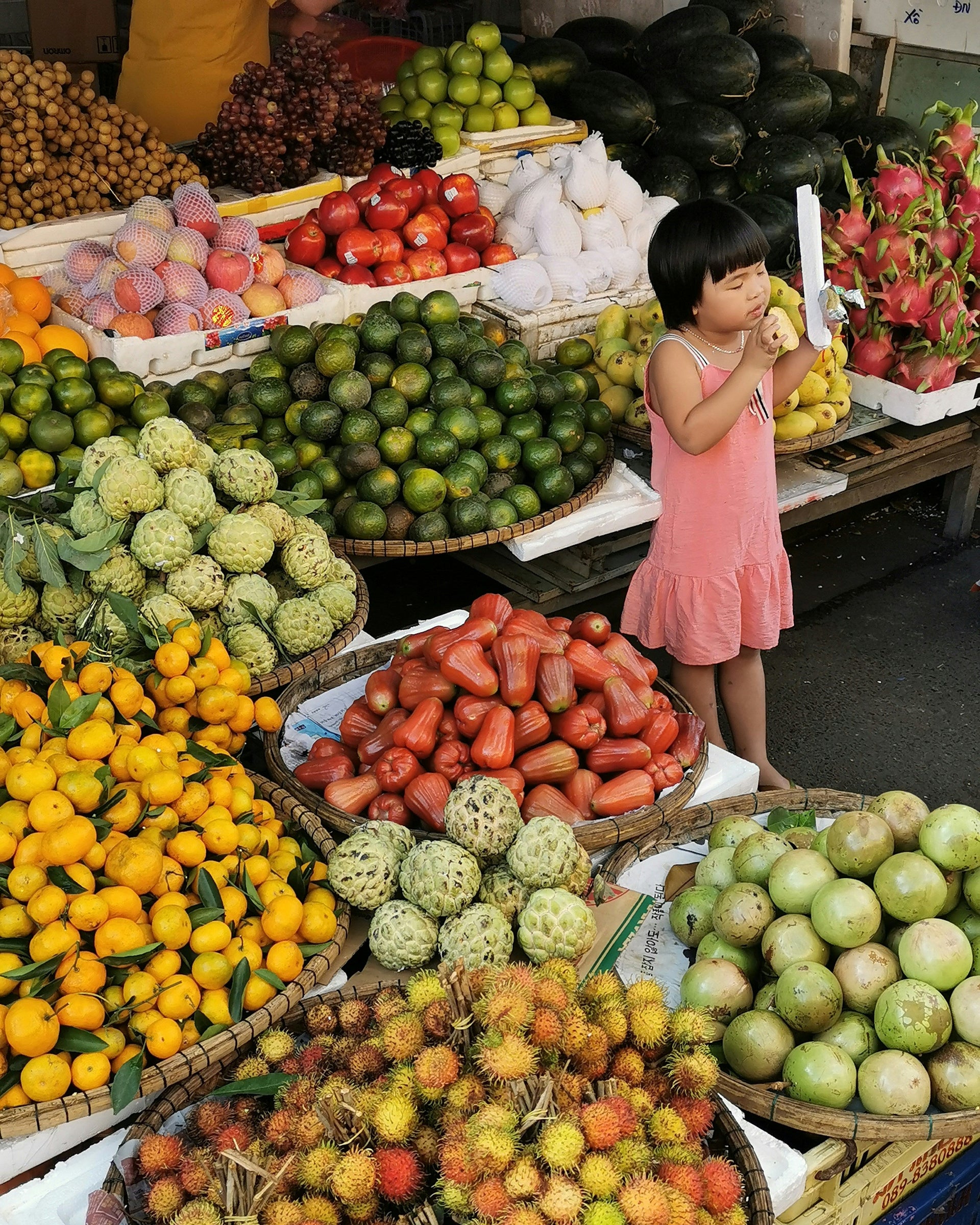 Child in a pink dress standing among various fruits and vegetables at an outdoor market.